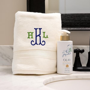 May include: A folded cream-colored hand towel featuring an embroidered monogram with the letters H, K, and L in green and blue thread, positioned next to a white Olay pump bottle containing 1 fluid ounce of product with gold accents.