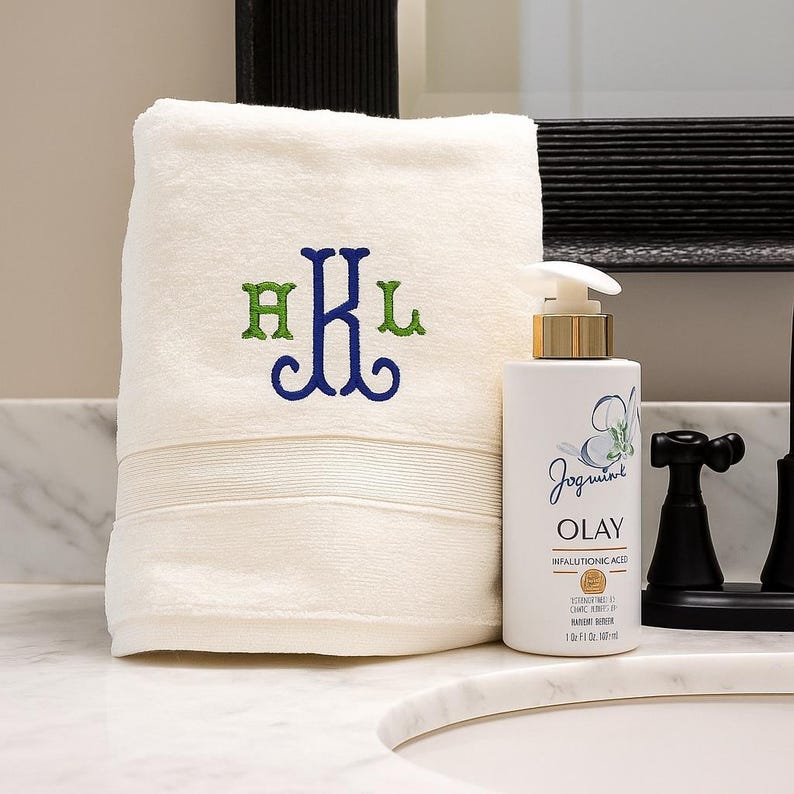 May include: A folded cream-colored hand towel featuring an embroidered monogram with the letters H, K, and L in green and blue thread, positioned next to a white Olay pump bottle containing 1 fluid ounce of product with gold accents.