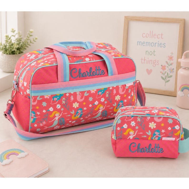 May include: A pink duffel bag and a matching toiletry bag, both featuring a repeating pattern of mermaids and flowers. Each bag has the name Charlotte embroidered in blue script. The duffel bag includes a rainbow-striped shoulder strap.