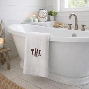 May include: A white bath towel featuring the embroidered monogram letters THK in a dark brown, vertical-striped font. The towel is draped over the edge of a white, oval-shaped freestanding bathtub with a metallic faucet and handles.