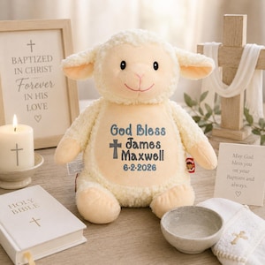 May include: A cream-colored plush lamb toy featuring embroidered text on its belly that reads God Bless James Maxwell 6-2-2026, accompanied by a cross symbol. The stuffed animal has soft, textured fur, black eyes, and a stitched pink mouth.