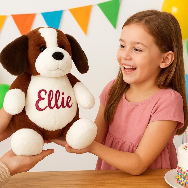 May include: A brown and white plush dog toy with the name "Ellie" embroidered in red script on its chest. The stuffed animal has floppy ears and white paws. The toy is being held by a person, and a young girl is smiling in the background.