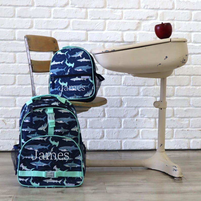 May include: Two navy blue backpacks with a shark pattern and the name "James" in white script. One backpack is on a wooden chair, and the other is on the floor. A vintage school desk and a red apple are in the background.