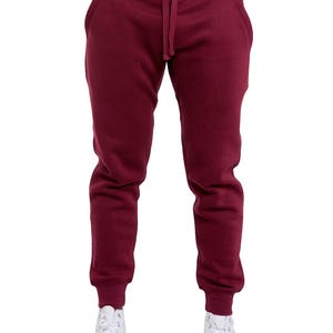 May include: Burgundy-colored sweatpants with a drawstring waist and elastic cuffs. The pants have side pockets and are paired with white high-top sneakers. The image is a full-length shot of the pants.