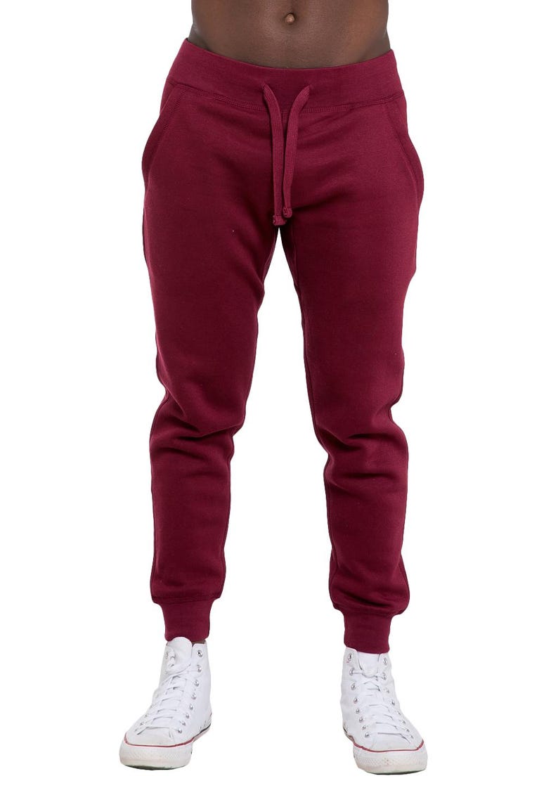 May include: Burgundy-colored sweatpants with a drawstring waist and elastic cuffs. The pants have side pockets and are paired with white high-top sneakers. The image is a full-length shot of the pants.