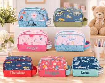 May include: A collection of seven rectangular zippered pouches in various colors and patterns, including floral, construction vehicles, ballerinas, castles, cars, mermaids, and sharks. Each pouch features a personalized name embroidered on the front.