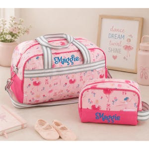 May include: A pink duffel bag and a matching toiletry bag, both featuring a ballerina print and the name Maggie embroidered in blue. The bags have grey and white striped handles and straps. A pair of light pink ballet slippers sits in front.