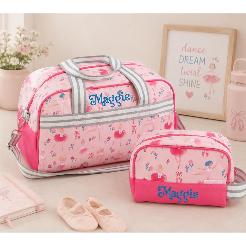 May include: A pink duffel bag and a matching toiletry bag, both featuring a ballerina print and the name Maggie embroidered in blue. The bags have grey and white striped handles and straps. A pair of light pink ballet slippers sits in front.