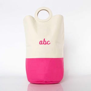 May include: A white canvas laundry basket with a pink bottom and a wooden handle. The basket has the letters "abc" embroidered on the front in pink.