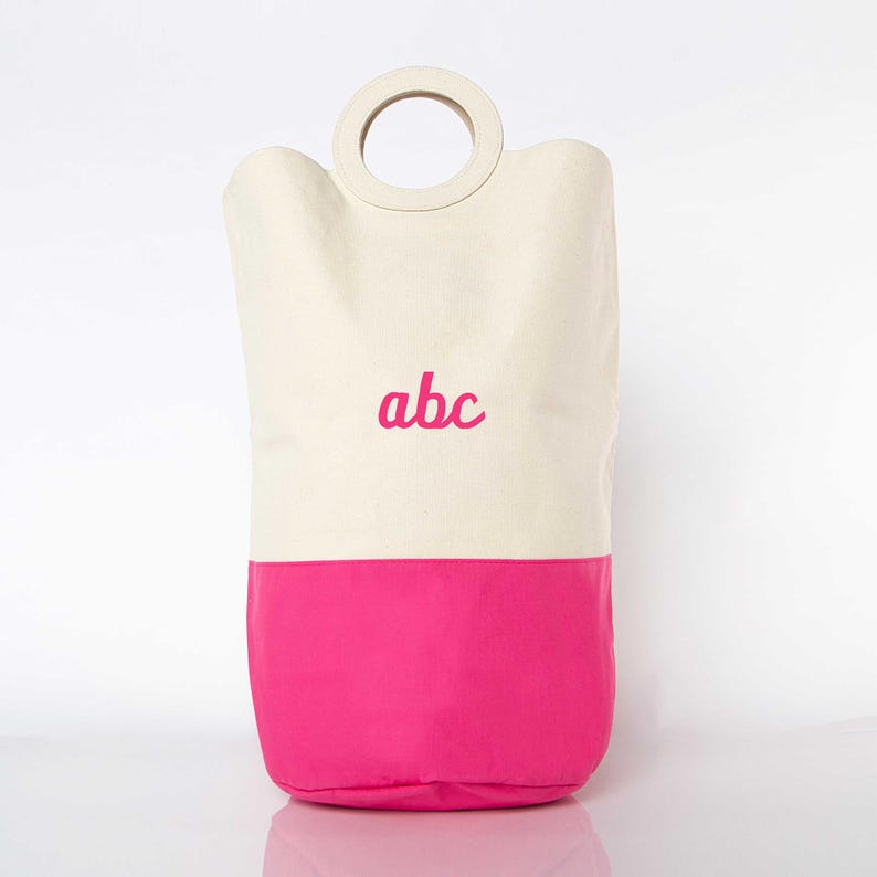 May include: A white canvas laundry basket with a pink bottom and a wooden handle. The basket has the letters "abc" embroidered on the front in pink.