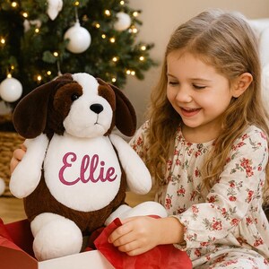 May include: A plush dog toy with brown and white fur, and floppy ears. The dog has the name "Ellie" embroidered in pink on its chest. The toy is sitting in a red gift box, with a Christmas tree in the background.
