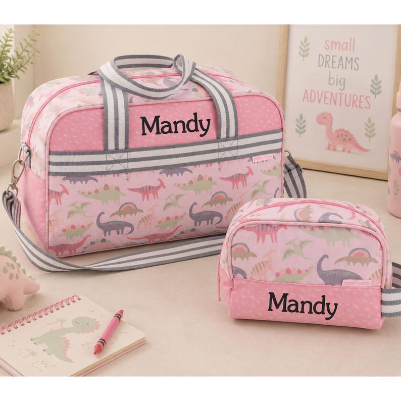 May include: A pink duffel bag and a matching smaller pouch, both featuring a dinosaur pattern and the name Mandy embroidered in black. The bags have grey and white striped handles and straps. The duffel bag has a rectangular shape with a zipper.