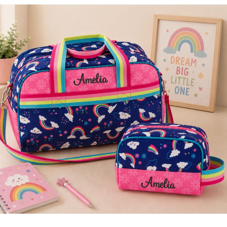 May include: A navy blue duffel bag and a matching toiletry bag, both featuring a colorful rainbow and cloud pattern, pink floral accents, and the name Amelia embroidered in black. The bags have bright rainbow-striped straps and trim.