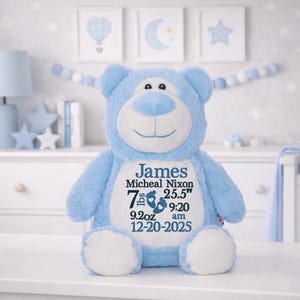 May include: A light blue plush teddy bear with white accents on the ears, muzzle, and feet. The chest features embroidered text: James, Micheal Nixon, 7 lbs 9.2 oz, 25.5 inches, 9:20 am, and 12-20-2025, along with two small blue baby footprint icons.