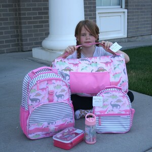May include: A set of pink and gray dinosaur-themed backpacks, a lunchbox, and a water bottle. The backpacks have a striped pattern and feature a pink and green dinosaur design. The lunchbox is pink and has a dinosaur design. The water bottle is clear with a pink lid and a dinosaur design.