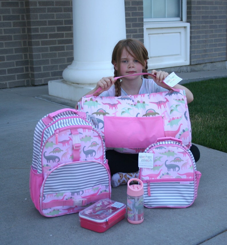 May include: A set of pink and gray dinosaur-themed backpacks, a lunchbox, and a water bottle. The backpacks have a striped pattern and feature a pink and green dinosaur design. The lunchbox is pink and has a dinosaur design. The water bottle is clear with a pink lid and a dinosaur design.