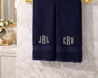 May include: Two navy blue bath towels hanging on a gold metal towel bar. Each towel features a horizontal band near the bottom and is embroidered with a three-letter monogram in a light gray serif font, reading JRL on one and CRR on the other.