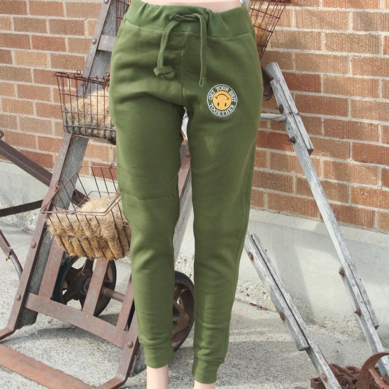 May include: Olive green sweatpants with a drawstring waist and a circular graphic on the left thigh. The graphic features a smiling face and the text "Get Your Shit Together". The pants are cuffed at the ankles.