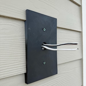 May include: A black rectangular electrical box mounted on a beige siding. Two screws secure the box to the siding. White, black, and gray wires extend from a hole in the box.