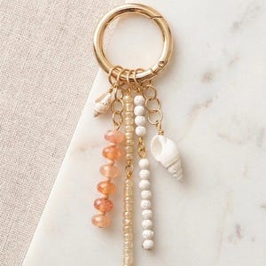 Peut inclure: Un porte-clés doré avec un fermoir rond, orné de divers breloques. Ceux-ci comprennent de petits coquillages, des perles de pierres précieuses orange, des perles transparentes et des perles blanches texturées, le tout suspendu à de fines chaînes dorées. Le fond est une surface en marbre blanc.