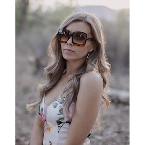 May include: Square-framed sunglasses with a tortoiseshell pattern on the outer frame and gradient lenses. The sunglasses are worn by a person with long, wavy blonde hair. The person is wearing a floral print dress.