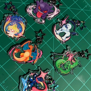 May include: Six acrylic keychains featuring colorful cartoon creatures, including a purple ghost, a yellow star-like figure, a white creature, an orange dragon, a green serpent, and a blue frog-like character, each with a black star-shaped clasp.