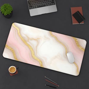 May include: A rectangular desk mat with a pink, white, and gold marble design. The mat has a black border and is shown with a laptop, mouse, and other office supplies. The design features flowing gold veins.