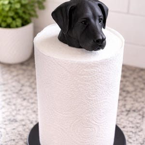 May include: A black dog head sculpture sits atop a roll of white paper towels. The paper towels are on a black circular base. The dog head is a detailed sculpture, and the paper towels have a textured pattern.