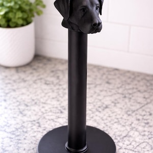 May include: Black metal paper towel holder featuring a sculpted dog head. The holder has a round base and a vertical pole, with the dog's head at the top. The design is simple and modern, suitable for a kitchen or bathroom.