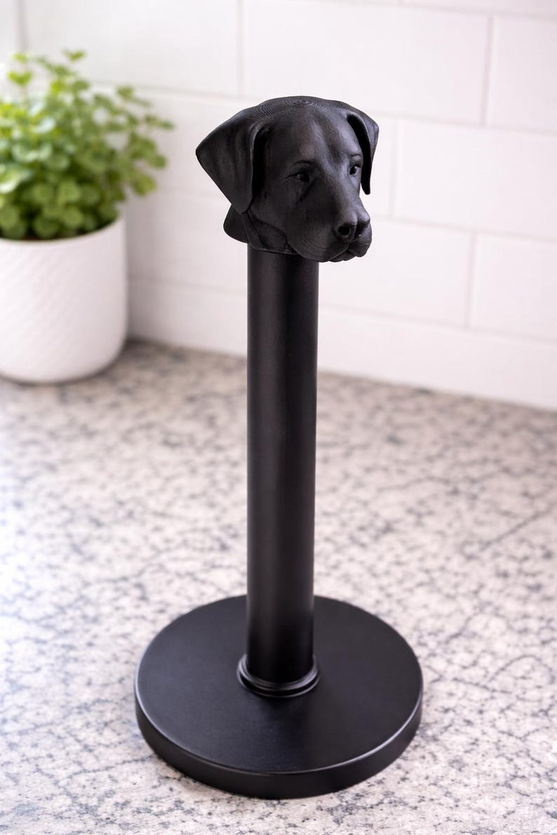 May include: Black metal paper towel holder featuring a sculpted dog head. The holder has a round base and a vertical pole, with the dog's head at the top. The design is simple and modern, suitable for a kitchen or bathroom.