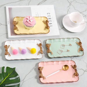 Op de afbeelding: Een verzameling decoratieve rechthoekige trays in verschillende pastelkleuren: geel, wit, mintgroen en roze. Elke tray heeft een geschulpte rand, strikvormige handvatten en is versierd met decoratieve items zoals een cupcake, pompons en sieraden. Een witte kop en schotel zijn ook aanwezig.