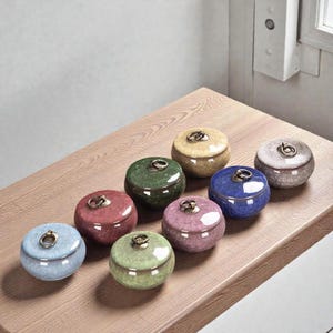 May include: A collection of eight small, round ceramic jars with lids. The jars are in various colors, including blue, green, red, and yellow, and have a glossy finish. Each jar has a small, gold-colored ring on top for opening.