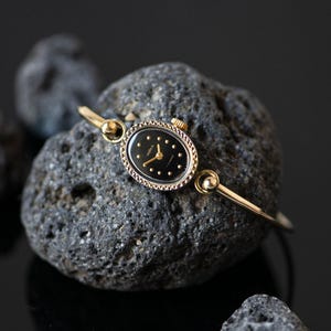 May include: A gold-tone bangle watch featuring an oval-shaped case with a black dial. The watch face displays gold-colored hour markers and thin gold hands. The bezel has a textured, decorative edge, and the band is a slim, polished gold-tone metal.