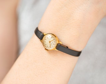 Op de afbeelding: Een gouden polshorloge met een witte wijzerplaat en een zwarte leren band. De wijzerplaat van het horloge heeft zwarte Romeinse cijfers als uurmarkeringen en een zwarte secondewijzer.