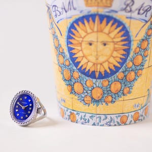 Puede incluir: Un anillo reloj plateado con esfera circular azul intenso, que presenta el texto "CHAIKA" y "17 JEWELS" en blanco. El bisel tiene un borde texturizado similar a una cuerda. Al lado se encuentra un recipiente cilíndrico con un sol amarillo.