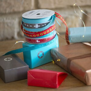 May include: A stack of four spools of decorative ribbon in light blue, grey with white patterns, red with yellow flowers, and solid red with white stitching, resting on a bright blue box with a star icon, surrounded by various wrapped gift boxes.