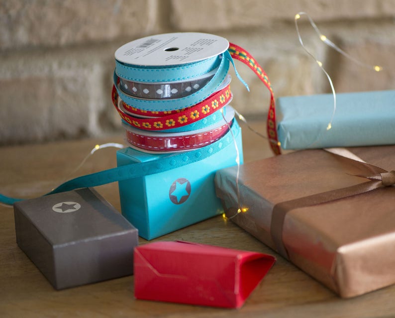 May include: A stack of four spools of decorative ribbon in light blue, grey with white patterns, red with yellow flowers, and solid red with white stitching, resting on a bright blue box with a star icon, surrounded by various wrapped gift boxes.