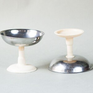 May include: Two vintage champagne coupe glasses. One glass is upright, showing a stainless steel bowl atop a white stem and base. The other glass is inverted, revealing the stainless steel base and white stem.