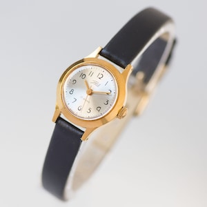 Peut inclure: Une montre-bracelet dor&eacute;e avec un bo&icirc;tier rond et un bracelet en cuir noir. Le cadran est argent&eacute; avec des chiffres arabes noirs, des aiguilles dor&eacute;es, et le texte Zaria et 17 jewels. Le bas du cadran porte la mention Made in USSR.
