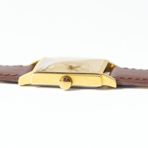 May include: A gold-toned wristwatch with a square face and a brown leather band. The watch has a simple design with a white dial and black hands.