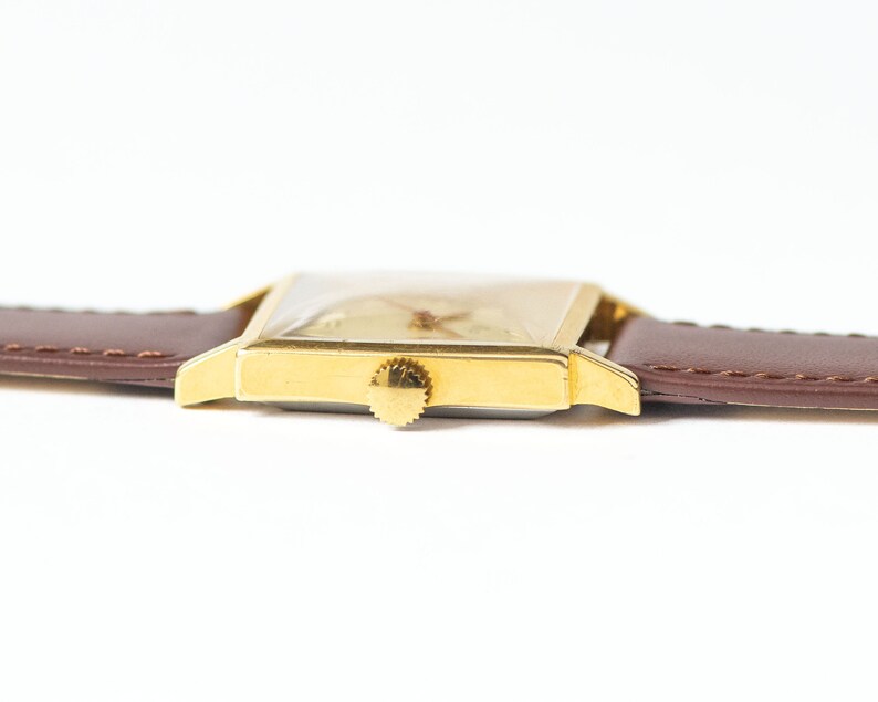 May include: A gold-toned wristwatch with a square face and a brown leather band. The watch has a simple design with a white dial and black hands.