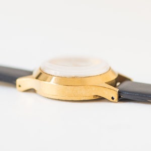 Peut inclure: Une montre-bracelet dor&eacute;e avec un bo&icirc;tier rond et un bracelet en cuir noir lisse. La montre est photographi&eacute;e sous un angle lat&eacute;ral, mettant en &eacute;vidence la finition m&eacute;tallique du bo&icirc;tier et les points de fixation du bracelet aux cornes.