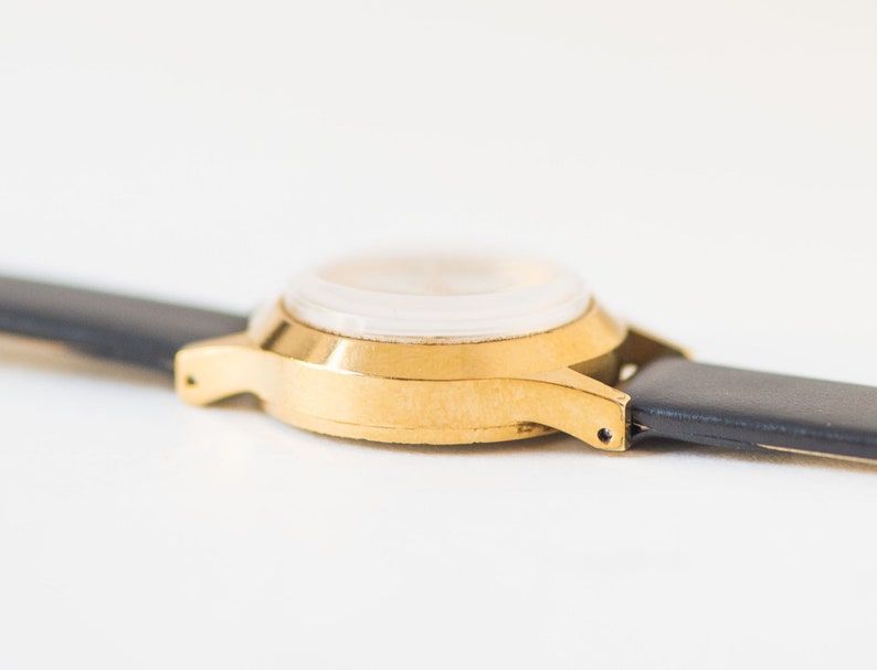 Peut inclure: Une montre-bracelet dor&eacute;e avec un bo&icirc;tier rond et un bracelet en cuir noir lisse. La montre est photographi&eacute;e sous un angle lat&eacute;ral, mettant en &eacute;vidence la finition m&eacute;tallique du bo&icirc;tier et les points de fixation du bracelet aux cornes.