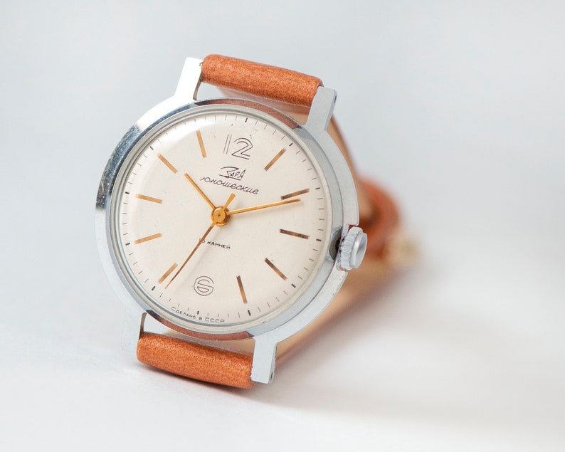 May include: A silver wristwatch with a white face and gold hands. The watch has a brown leather band and the words "ЧacOBHИK" and "СДЕЛАНО В СССР" are printed on the face.