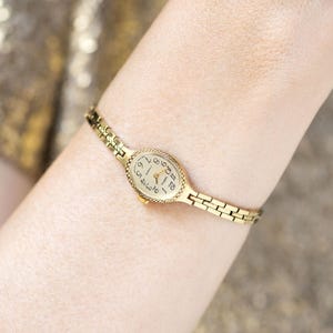 May include: A gold-tone wristwatch with an oval-shaped case and a textured bezel. The watch features a cream-colored dial with black Arabic numerals and thin gold hands. It is attached to a delicate, slender link bracelet with a polished finish.