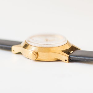 Peut inclure: Une montre-bracelet dor&eacute;e dot&eacute;e d'un bo&icirc;tier rond avec une couronne de remontage lat&eacute;rale. La montre est fix&eacute;e &agrave; un bracelet en cuir noir lisse qui s'&eacute;tend horizontalement depuis les cornes de chaque c&ocirc;t&eacute; du bo&icirc;tier m&eacute;tallique.
