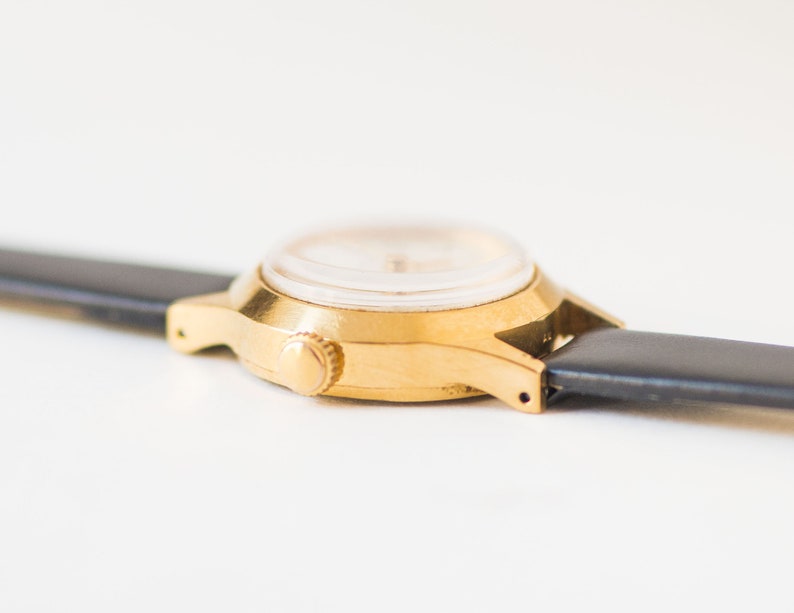 Peut inclure: Une montre-bracelet dor&eacute;e dot&eacute;e d'un bo&icirc;tier rond avec une couronne de remontage lat&eacute;rale. La montre est fix&eacute;e &agrave; un bracelet en cuir noir lisse qui s'&eacute;tend horizontalement depuis les cornes de chaque c&ocirc;t&eacute; du bo&icirc;tier m&eacute;tallique.