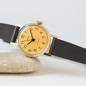 Könnte beinhalten: Eine goldfarbene Armbanduhr mit einem schwarzen Lederarmband. Das Zifferblatt der Uhr ist gelb mit schwarzen Zahlen und Zeigern. Das Zifferblatt der Uhr trägt außerdem den Text "ВАРЯ" und "21 КАНЕЛЬ".