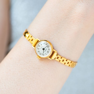 May include: A gold wristwatch with a white face and black numbers. The watch has a gold metal band.