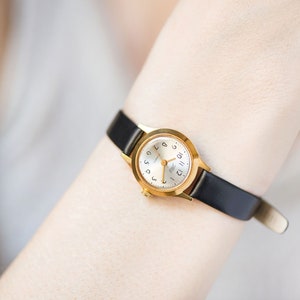 Peut inclure: Une montre-bracelet dor&eacute;e dot&eacute;e d'un bo&icirc;tier rond avec un cadran blanc, des chiffres arabes noirs pour les heures et de fines aiguilles dor&eacute;es. La montre est fix&eacute;e &agrave; un bracelet en cuir noir fin et lisse avec une boucle dor&eacute;e et une couronne.